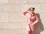 Young beautiful smiling hipster woman in trendy summer pink overall. Carefree teen model posing in the street near wall at sunset. Positive female outdoors in sunglasses. Cheerful and happy.