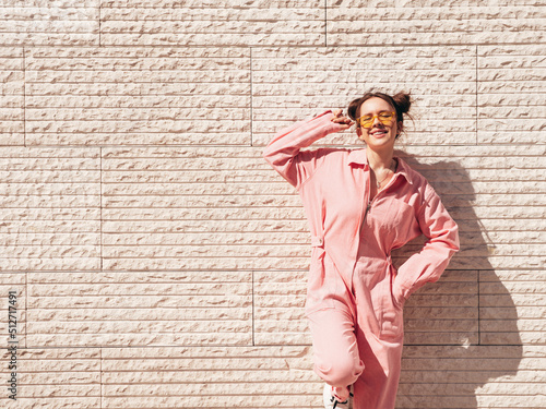 Young beautiful smiling hipster woman in trendy summer pink overall. Carefree teen model posing in the street near wall at sunset. Positive female outdoors in sunglasses. Cheerful and happy.