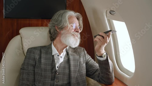 Senior male millionaire talking on the speakerphone or recording a voice message on his smartphone while flying on a private jet. Rich man talking on mobile phone while flying at first class airplane photo