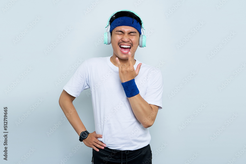 Crazy young Asian man sporty fitness trainer instructor in blue headband and white t-shirt. Listen music with headphones, Depicting heavy metal rock sign isolated over white background Crazy young Asian man sporty fitness trainer instructor in blue headband and white t-shirt. Listen music with headphones, Depicting heavy metal rock sign isolated over white background