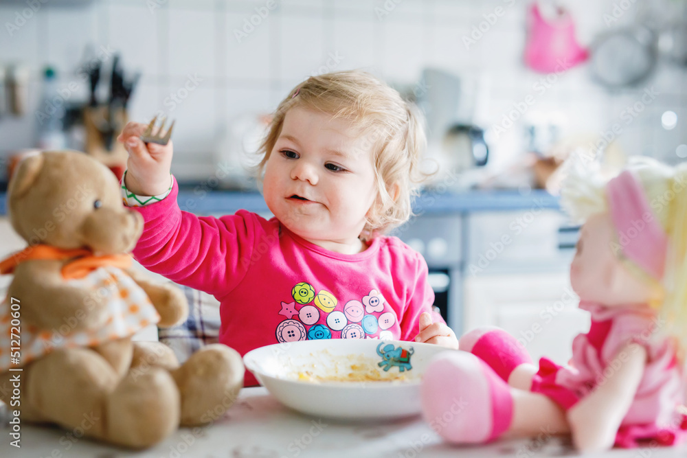 Fototapeta premium Adorable baby girl eating from fork vegetables and pasta. food, child, feeding and development concept. Cute toddler, daughter with spoon sitting in highchair and learning to eat by itself.