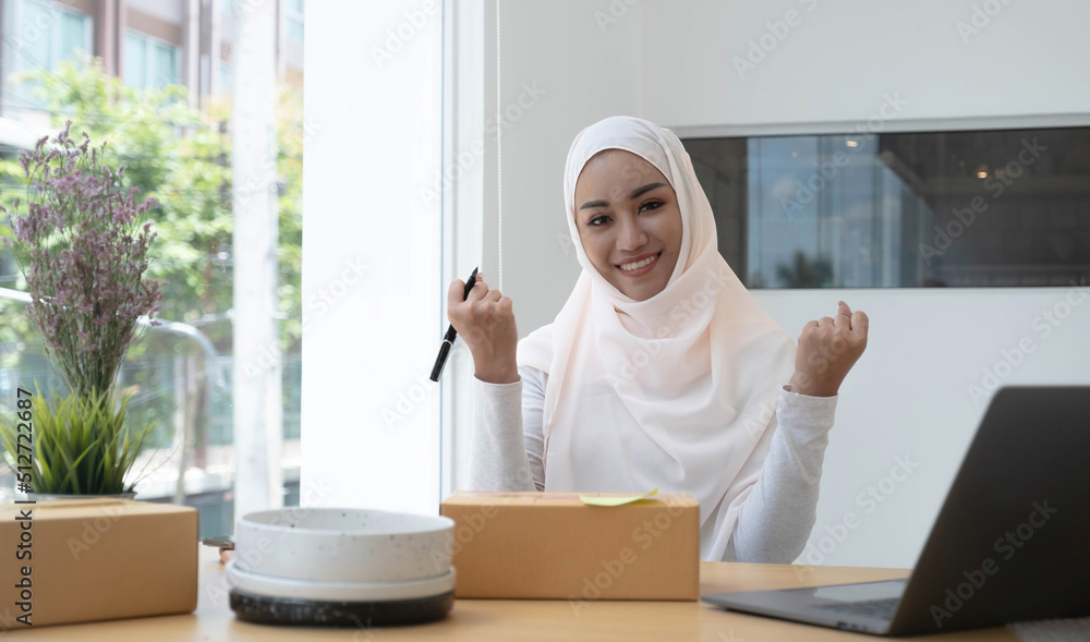 Portrait of a cute smiling Muslim woman wearing a light pink hijab sitting on an office chair. Looking at the camera is delighted while being able to sell online and the background of the box. Portrait of a cute smiling Muslim woman wearing a light pink hijab sitting on an office chair. Looking at the camera is delighted while being able to sell online and the background of the box.