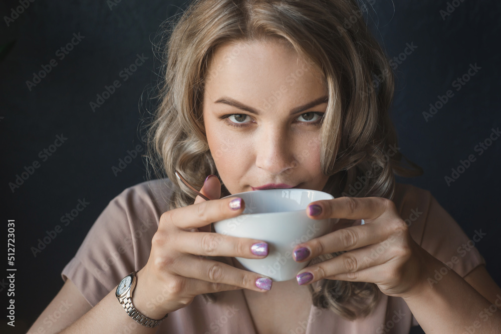 A young chubby woman in drinks coffee from a white cup on a dark background. serious look at the camera