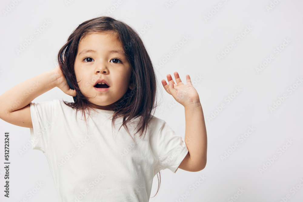 a little girl of preschool age, sitting on a white background in a white T-shirt, enthusiastically talking on the phone, turning half-side to the camera. The topic of the conversation on the phone a little girl of preschool age, sitting on a white background in a white T-shirt, enthusiastically talking on the phone, turning half-side to the camera. The topic of the conversation on the phone