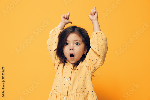 a cute, joyful preschool girl stands on an orange background and dances happily in a beautiful summer dress with her hands up