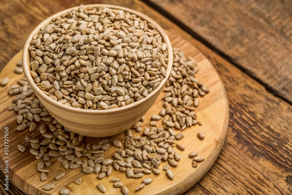 Bowl with peeled sunflower seeds and board on wooden background, closeup