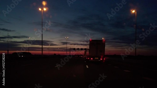 POV Driving car on highway late at night with cars and trucks with street lights and guardrails along the road after sunset. Beautiful colorful sky with orange, red and blue sky. Camera on dashboard
