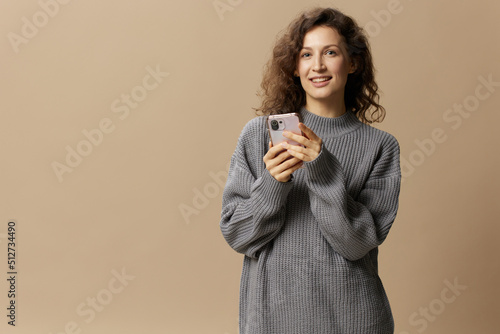 Happy curly beautiful lady in gray casual sweater chatting with boyfriend looks at camera posing isolated on beige pastel background. Social media, network, distance communication concept. Copy space