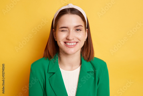 Indoor shot of playful childish woman wearing green jacket posing isolated over yellow background, looking at camera with toothy smile and winking, flirting with boyfriend. photo