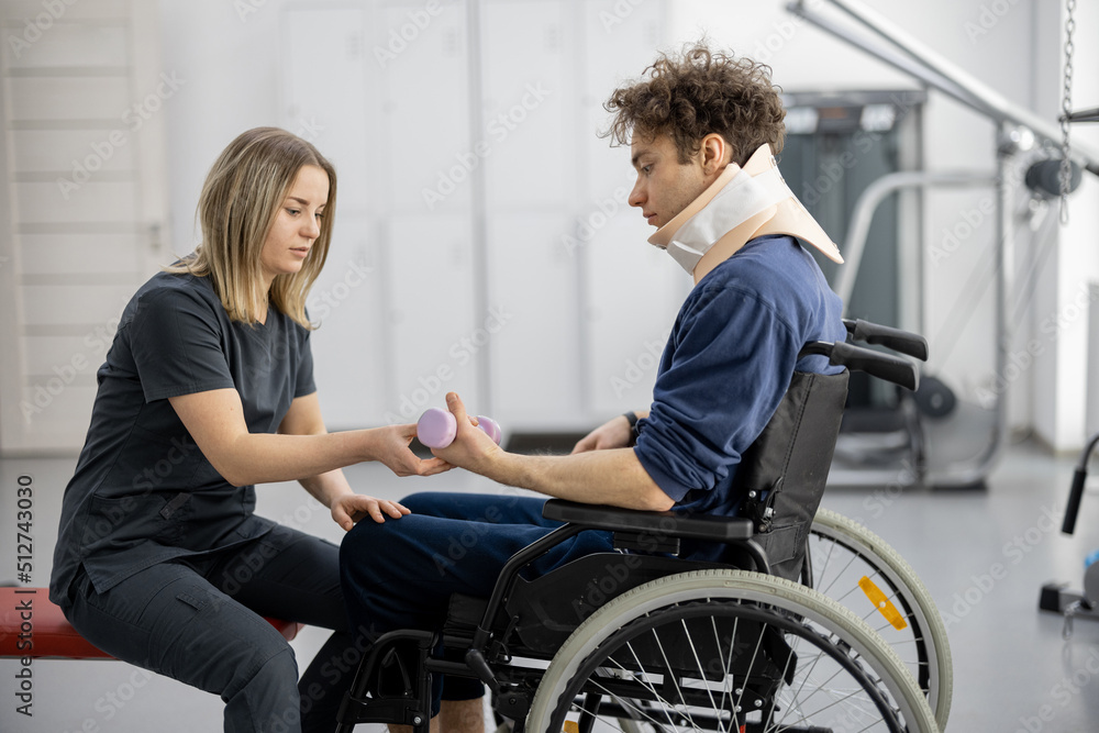 Rehabilitation specialist helps a guy to do exercises for recovery from injury, who is sitting in a wheelchair with a corset around his neck. Concept of physical therapy for people with disabilities Rehabilitation specialist helps a guy to do exercises for recovery from injury, who is sitting in a wheelchair with a corset around his neck. Concept of physical therapy for people with disabilities