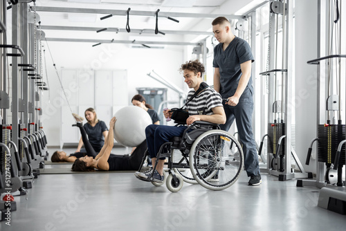 Leinwand Poster People exercising at rehabilitation center, rehabilitologist walking with guy in a wheelchair