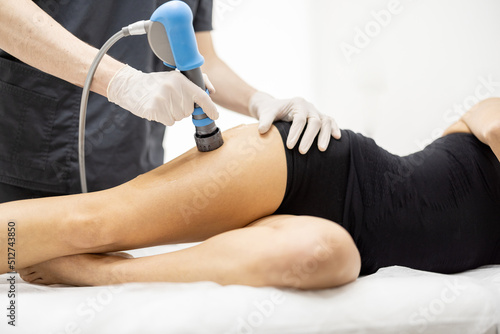 Doctor applies shock wave therapy with special medical equipment on women's thigh muscle at medical office. Concept of non-invasive technology for treating pain in musculoskeletal system