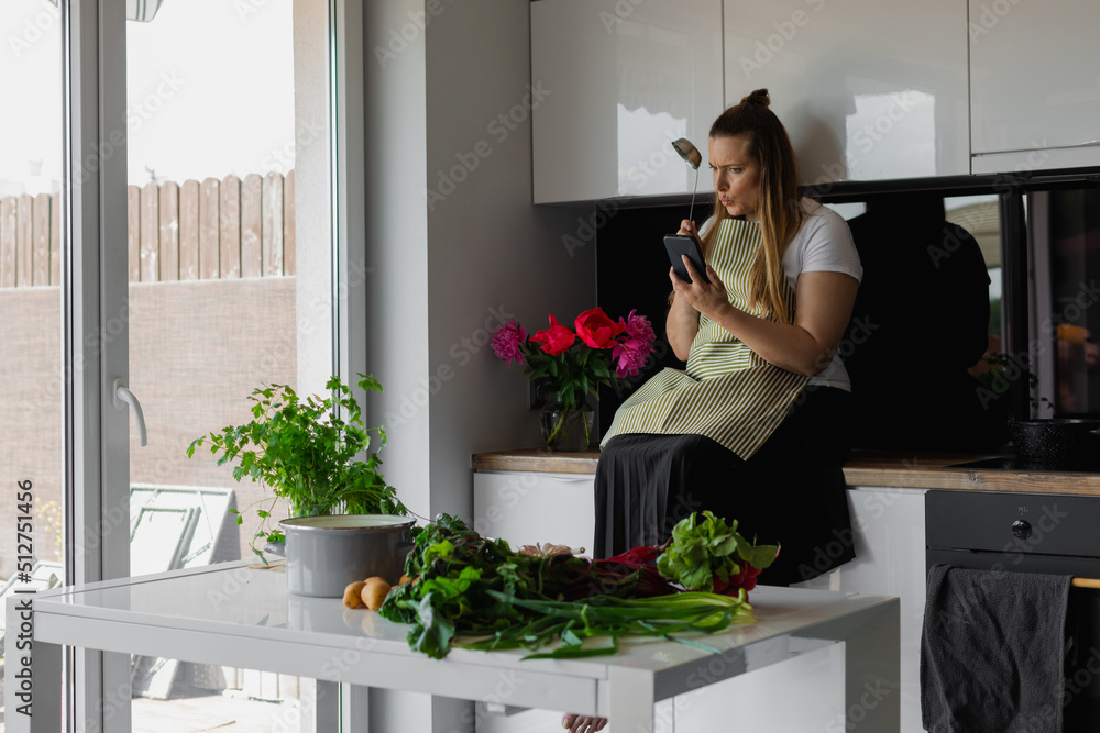 Inspired and passionate, plump, plus size blond woman with ladle sit on kitchen drawer with smartphone. Fresh vegetables Inspired and passionate, plump, plus size blond woman with ladle sit on kitchen drawer with smartphone. Fresh vegetables