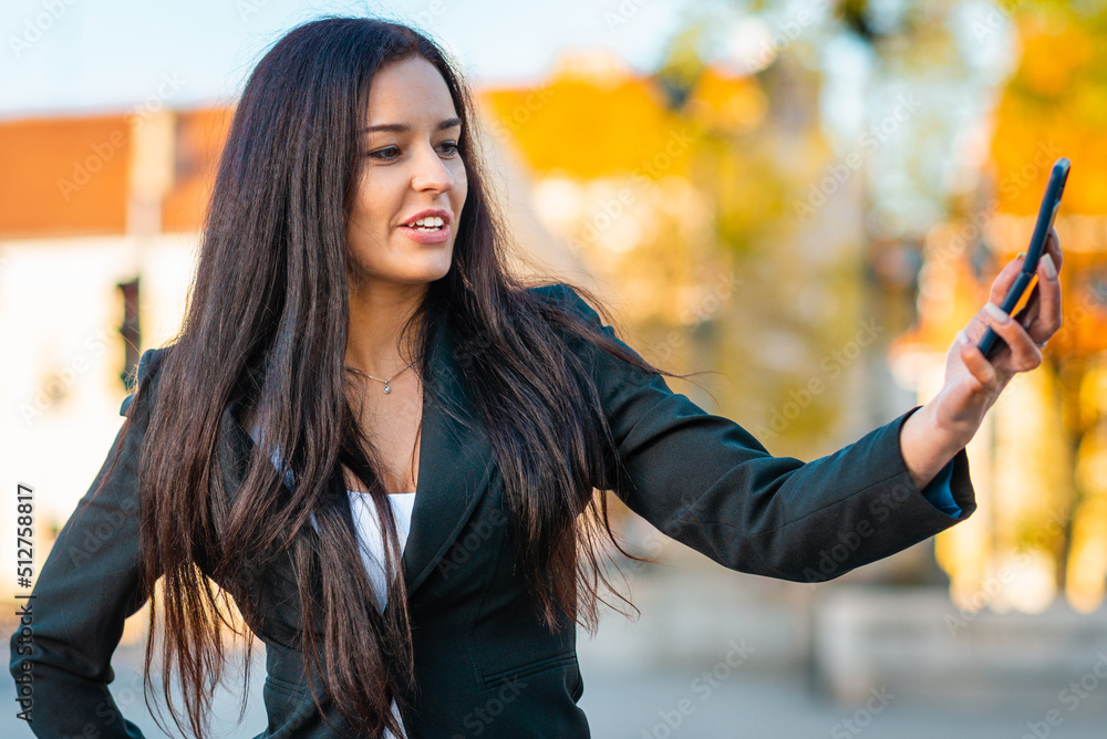 Portrait of a young beautiful business woman in a suit.Busineswoman walking through the city, video call, selfie, holding a phone.New generation business, communication.Blurred summer sunset.Close-up.