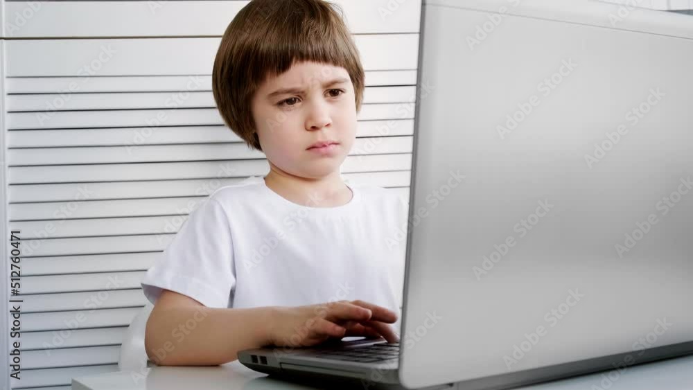 A focused and attentive child sits and does a task on a laptop. Close-up. 5 year old boy solves a test by a distance online education program. Smart kid. White room, furniture, t-shirt. Copy space. A focused and attentive child sits and does a task on a laptop. Close-up. 5 year old boy solves a test by a distance online education program. Smart kid. White room, furniture, t-shirt. Copy space.