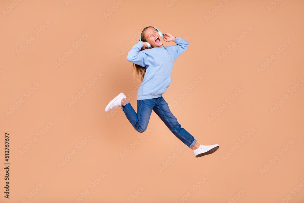 Full size photo of satisfied girl jump hands touch headphones scream have fun isolated on beige color background Full size photo of satisfied girl jump hands touch headphones scream have fun isolated on beige color background