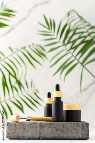 Concrete black podium for cosmetic products close-up among tropical leaves against the background of a marble white wall, bottle with a pipette and a jar of cream and a massage roller for the face