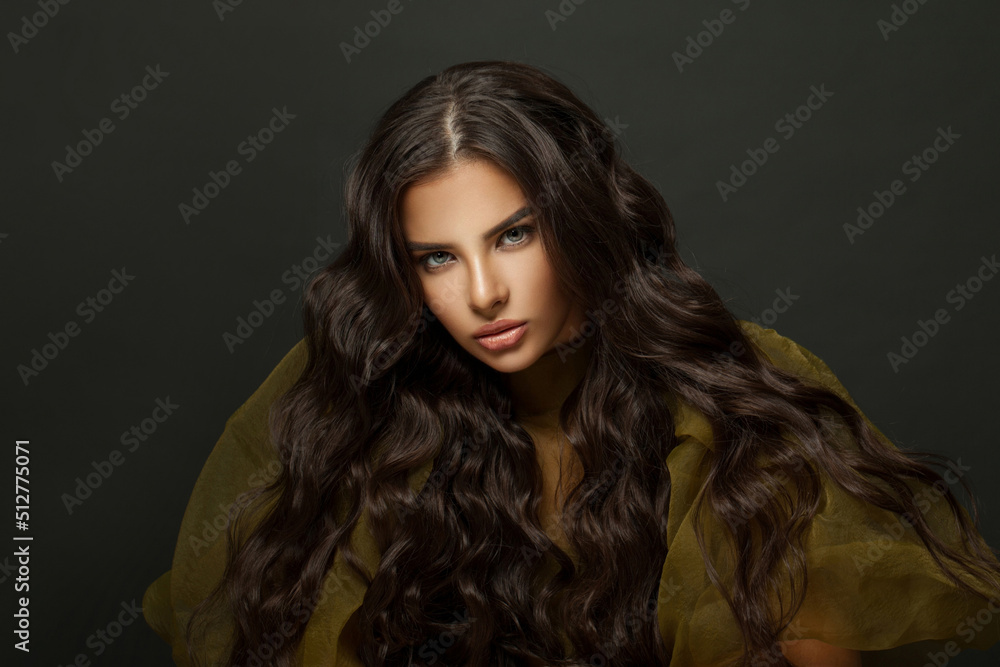 Fashion beauty portrait of glamorous woman with long dark wavy hair looking at camera against black studio wall background
