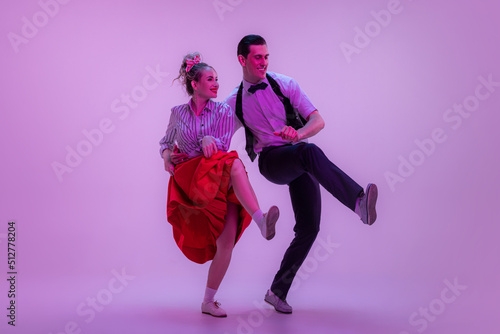 Studio shot of young man and woman in vintage retro style outfits dancing lindy hop isolated on lilac color background in neon light. photo
