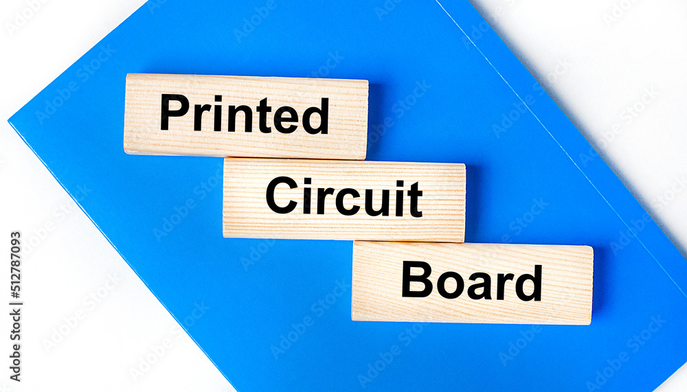 There is a blue notebook on a light gray background. Above are three wooden blocks with the words PCB Printed Circuit Board There is a blue notebook on a light gray background. Above are three wooden blocks with the words PCB Printed Circuit Board