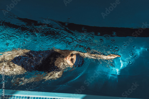 High angle of professional sportswoman in goggles and swimsuit swimming in backstroke or back crawl style during training in pool on sunny day  photo
