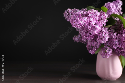 Beautiful lilac flowers in vase on wooden table against black background. Space for text