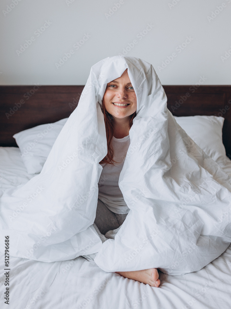 Beautiful young happy woman wrapped in soft white blanket sitting at home. Winter atmosphere. Millennial Caucasian girl wake up in cozy bed at home. Beautiful young happy woman wrapped in soft white blanket sitting at home. Winter atmosphere. Millennial Caucasian girl wake up in cozy bed at home.