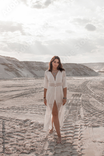 rustic style. close up brunette girl in white boho style dress and silver jewelry is walking casual and looking apart on the sand career background in desert at sunset. lifestyle concept  free space