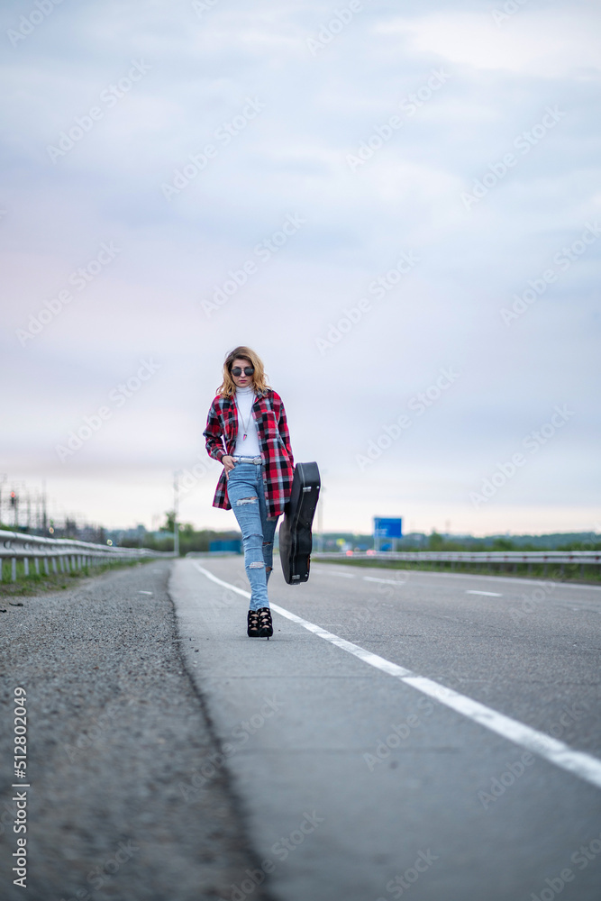 Cool woman with curly brown hair, in an unbuttoned plaid shirt, torn jeans, a white turtleneck, high heels, sitting on the road fence with electric guitar. Hitchhiker or traveler musician