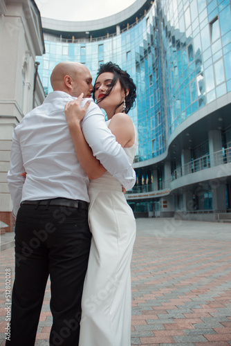 romantic wedding couple. groom with happy bride in hugs are standing close and smiling happy with kiss on the blue glass street building background at city. wedding concept, free space