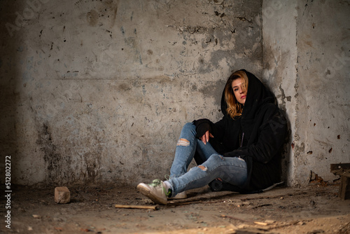 A mysterious goth woman in a black robe with a hood, torn jeans, sits thoughtfully on the dirty floor in an abandoned and destroyed building. Mysticism, post-apocalypse, depressed loneliness, witch