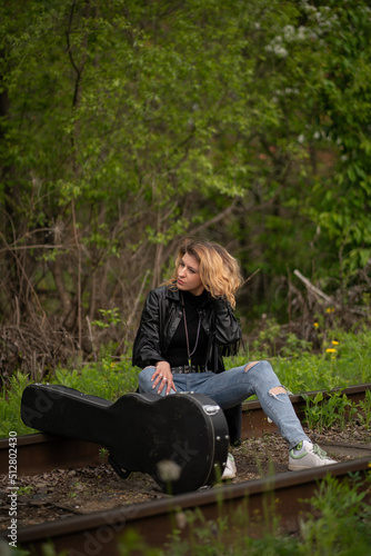 a brutal stylish female rock musician in a black raincoat and ripped jeans sits on the railway tracks in the forest and carries a guitar case. Traveler musician or adventurer