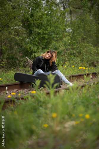 a brutal stylish female rock musician in a black raincoat and ripped jeans sits on the railway tracks in the forest among the grass and carries a guitar case. Traveler musician or adventurer