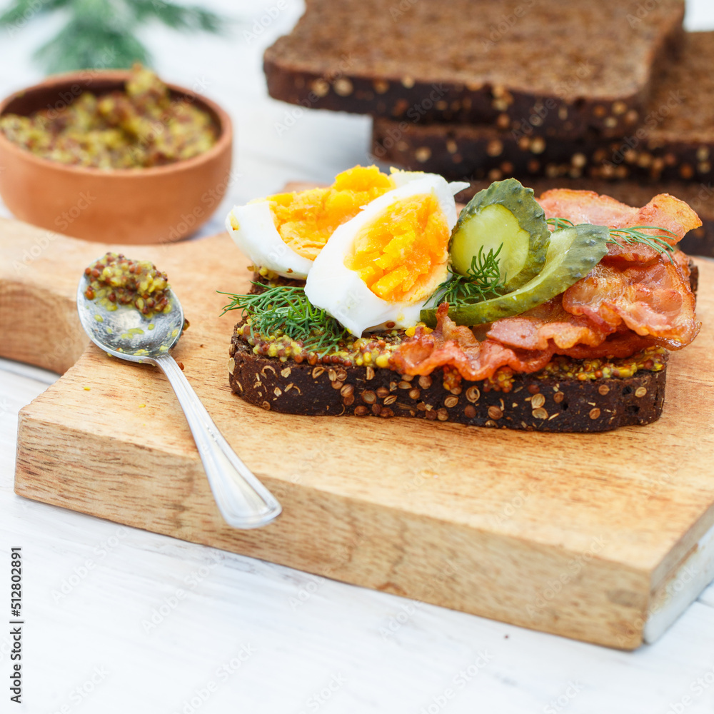 Delicious sandwich of rye bread with coriander with fried bacon, pickled cucumber, egg, dill and grainy mustard. A gourmet snack on a wooden board. Selective focus Delicious sandwich of rye bread with coriander with fried bacon, pickled cucumber, egg, dill and grainy mustard. A gourmet snack on a wooden board. Selective focus
