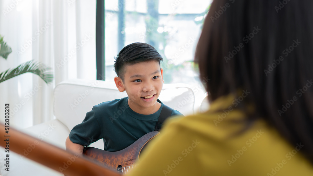 Asian boy playing guitar with mom in the living room for teaching him son play guitar, feel appreciated and encouraged. Concept of a happy family, learning and fun lifestyle, love family ties Asian boy playing guitar with mom in the living room for teaching him son play guitar, feel appreciated and encouraged. Concept of a happy family, learning and fun lifestyle, love family ties