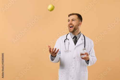 The male nutritionist doctor with stethoscope smiling and holding water and apple on beige background, diet plan concept photo