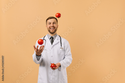 The male nutritionist doctor with stethoscope smiling and juggling tomatoes on beige background, diet plan concept photo