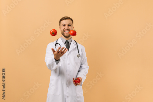 The male nutritionist doctor with stethoscope smiling and juggling tomatoes on beige background, diet plan concept