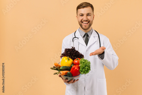 The happy male doctor nutritionist with stethoscope shows fresh vegetables on beige background, diet plan concept photo