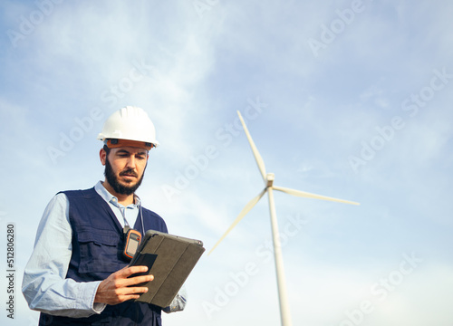 Young male engineer with a protective helmet works with a tablet in a field of windmills