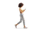 Full length profile shot of a young woman with a curly hair typing on a smartphone and walking