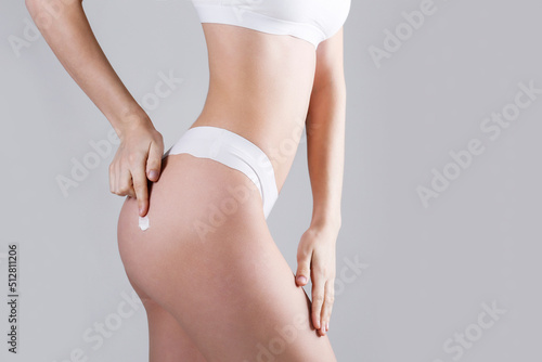 Studio shot of fit woman over isolated white background wearing only lingerie, applying moisturizing cream. Female in white underwear using a skincare product. Copy space for text, close up.