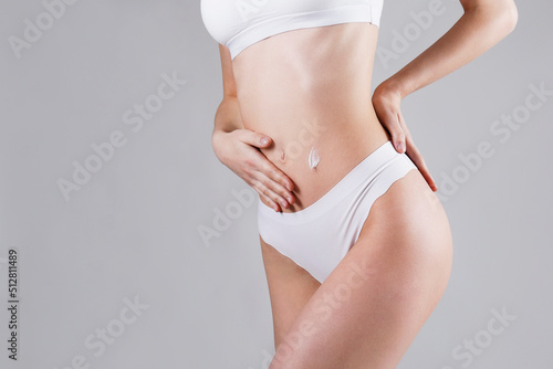 Studio shot of fit woman over isolated white background wearing only lingerie, applying moisturizing cream to her stomach. Female in white underwear using a skincare product. Copy space, close up.