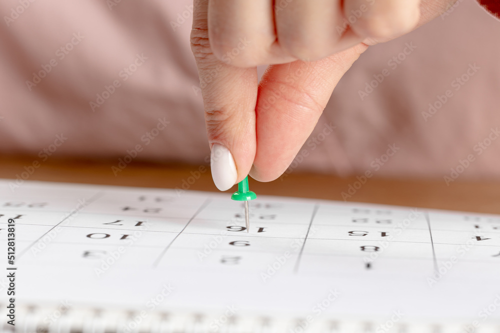 a female hand with a beautiful manicure holds a green pushpin to mark the date on the calendar a female hand with a beautiful manicure holds a green pushpin to mark the date on the calendar