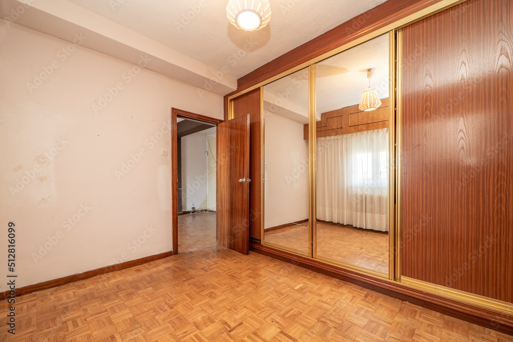 Empty bedroom with built-in wardrobe with mirrors and wooden sliding doors, golden aluminum, parquet floor and window with white curtains