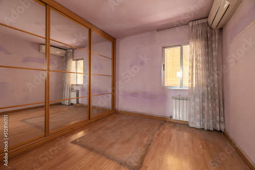 Empty bedroom with wardrobe with mirrored sliding doors, window with flower curtains, air conditioning and light oak parquet floor