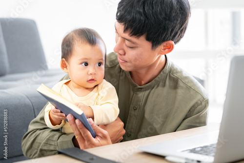 A baby daughter and an Asian father are reading a book together at home. and father has to work with