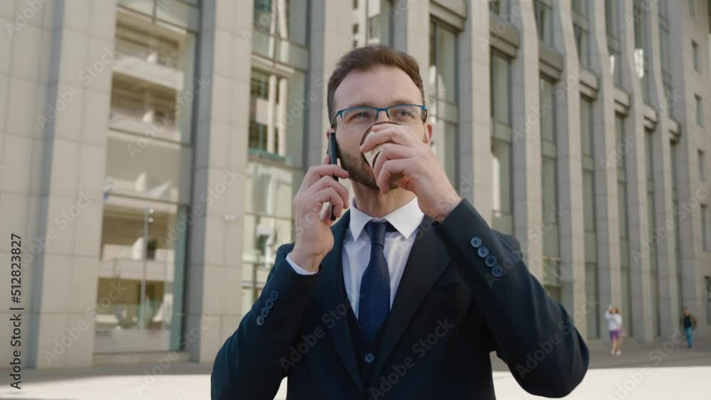 Handsome bearded businessman in formal suit talking mobile phone and drinking coffee while standing outside at downtown near office buildings Handsome bearded businessman in formal suit talking mobile phone and drinking coffee while standing outside at downtown near office buildings