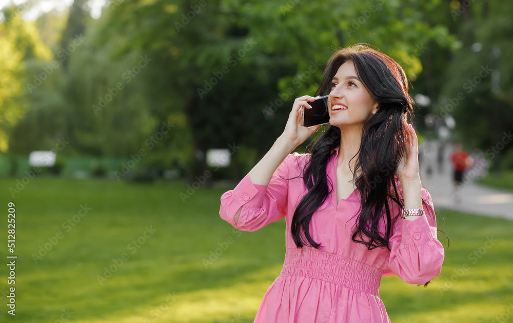 Close up portrait of a young brunette with wavy hair dressed in a pink dress walking in the park talking on a cell phone. Close up portrait of a young brunette with wavy hair dressed in a pink dress walking in the park talking on a cell phone.