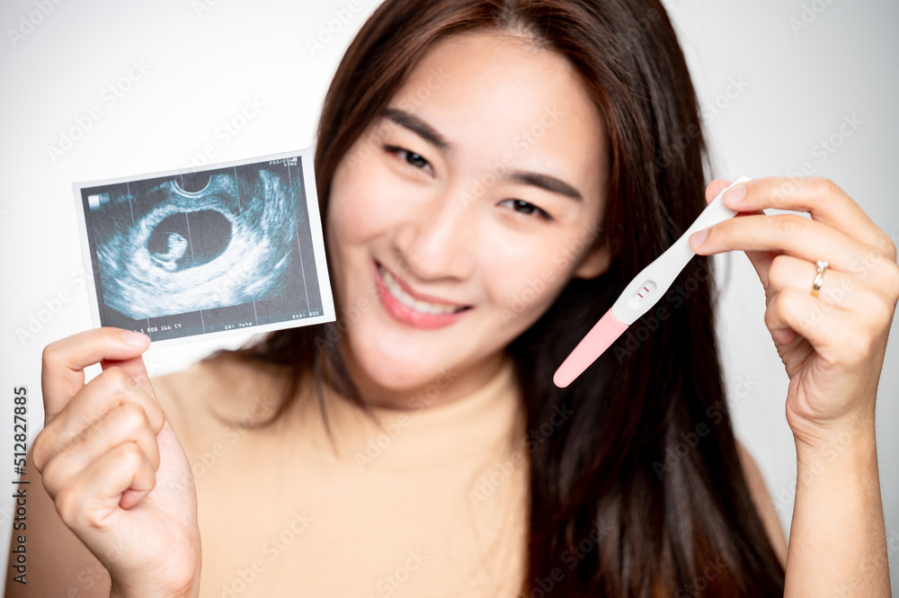 Asian woman holding ultrasound scan and pregnancy test showing a positive. Concept successful treatment IUI Intra – Uterine Insemination and ICSI. Asian woman holding ultrasound scan and pregnancy test showing a positive. Concept successful treatment IUI Intra – Uterine Insemination and ICSI.